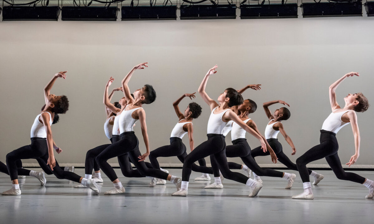 A group of young male dancers on stage wear white leotards with black leggings and white ballet shoes. They strike a lunging pose in unison, holding one arm curved in front of them and looking upwards. They are performing Eliot Feld&#039;s &quot;Hello Fancy.&quot;